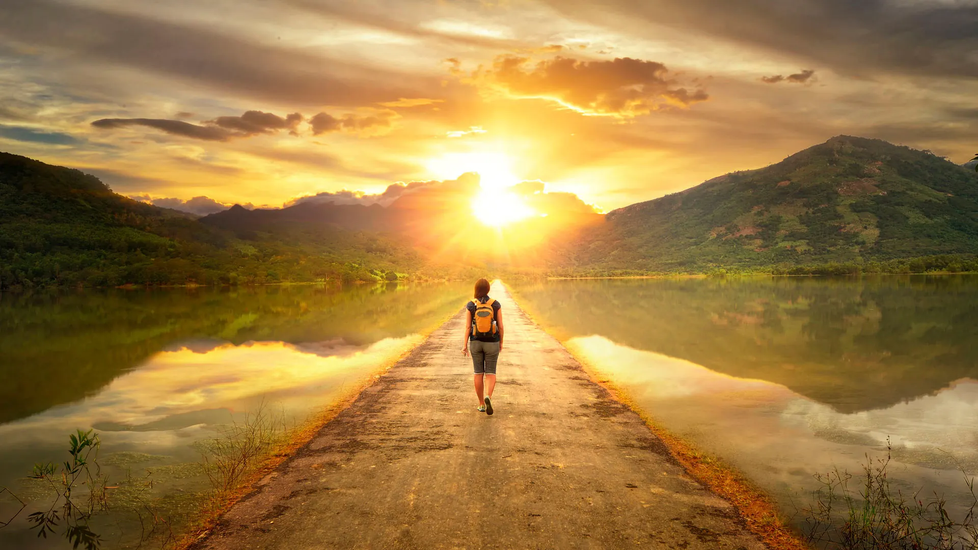 Eine Wanderin geht auf einem Damm zwischen zwei Seen dem Sonnenuntergang entgegen